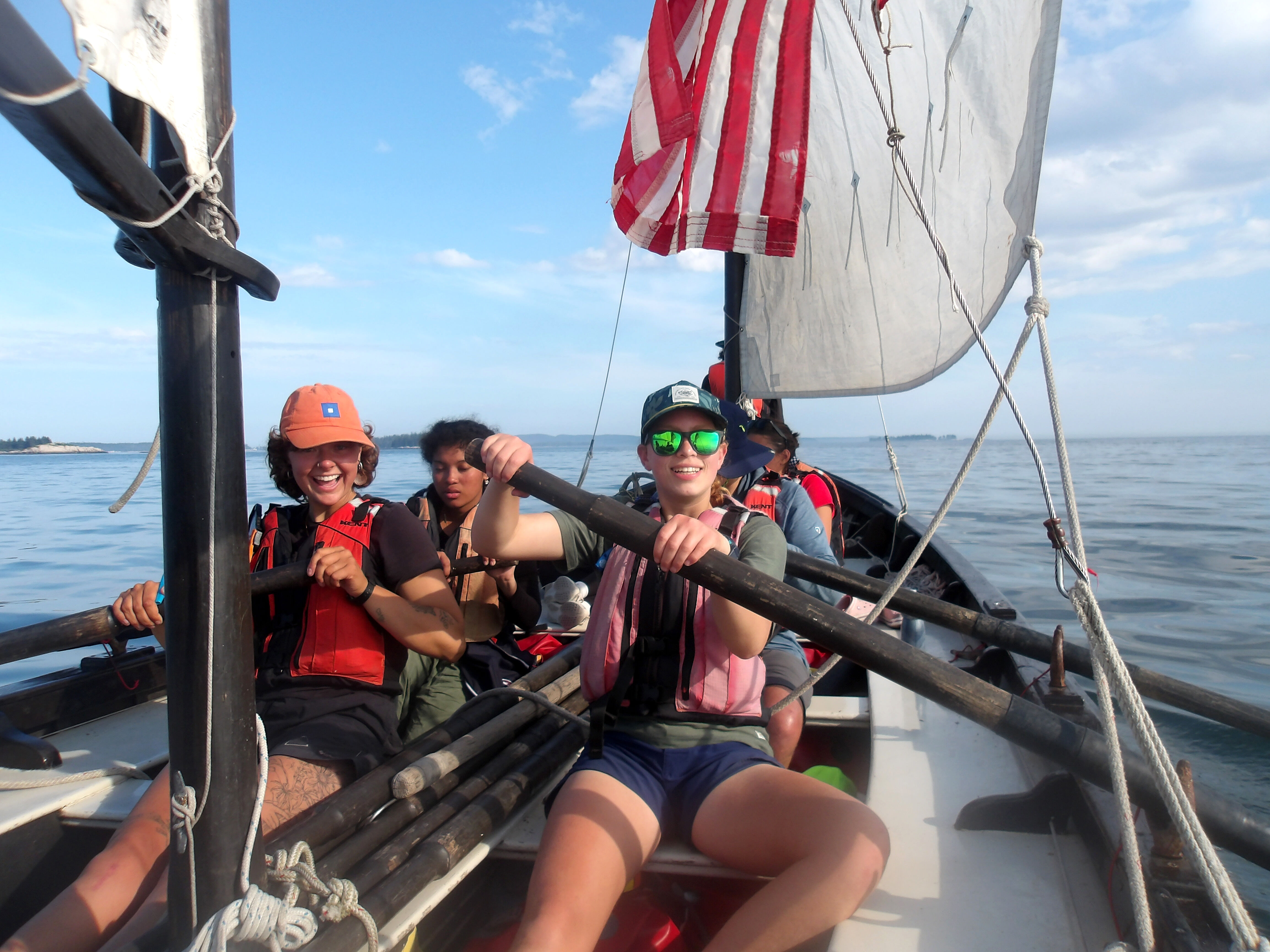 The image shows three people on a sailboat. The person in the foreground is wearing a pink life vest, sunglasses, and a hat. They are holding an oar and smiling. Behind them, another person is also holding an oar and wearing a life vest. A third person is visible in the background. The sailboat has an American flag attached to it, and the sky is blue with some clouds.